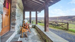 Un bâtiment rustique en pierre, doté d’une porte en bois et d’un porche abrité qui offre une vue sur un paysage rural.