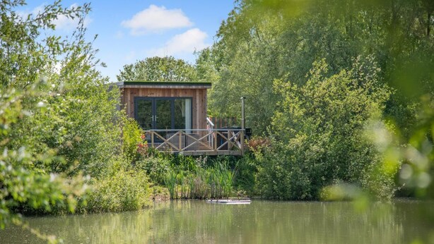 A wooden cabin with a glass door, surrounded by lush greenery and a body of water. - 1 Bed in Newport (oc-f30846) (Newport)