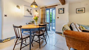 A dining area with a wooden table and chairs, a pendant light, and a vase of flowers. There is a sofa with cushions and a painting on the wall. - 2 Bed in Pentre Bach (oc-b30875) (Pentre-bach)