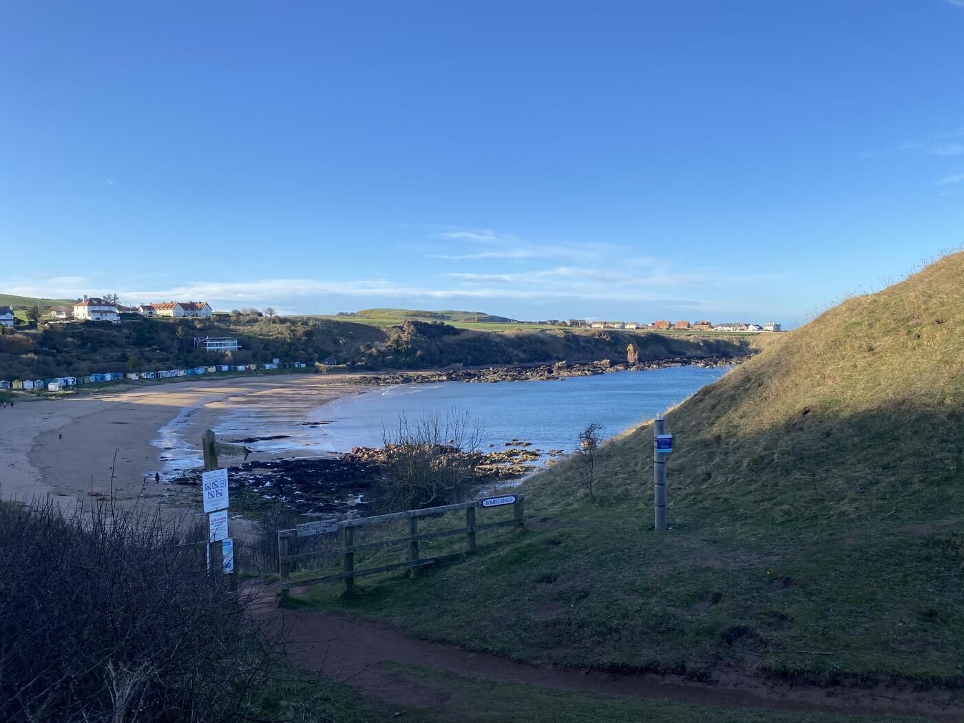 A coastal landscape with a sandy beach, a grassy hill, and a clear blue sky.