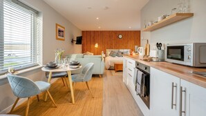 A compact, modern kitchen with a microwave, oven, and wooden countertop. There is a dining area with a table and chairs, and a bedroom with a bed and a wooden panel wall. - 1 Bed in Coldingham (oc-c30814) (Coldingham)