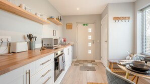 A modern kitchen with wooden shelves, a microwave, toaster, and a dining area with a table and chairs. - 1 Bed in Coldingham (oc-c30814) (Coldingham)