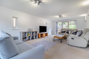 A modern living room with a grey sofa, a beige sofa, a flat-screen TV, a wooden entertainment unit, and a large window with a view of greenery. - 4 Bed in Caistor (oc-t30831) (Caistor)