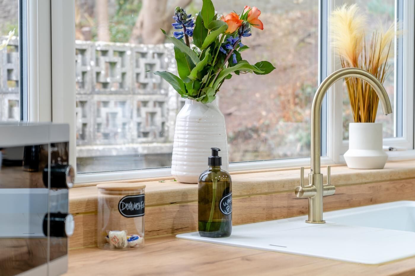 A kitchen counter with a window, a vase with flowers, a bottle of dish soap, and a jar of spices.