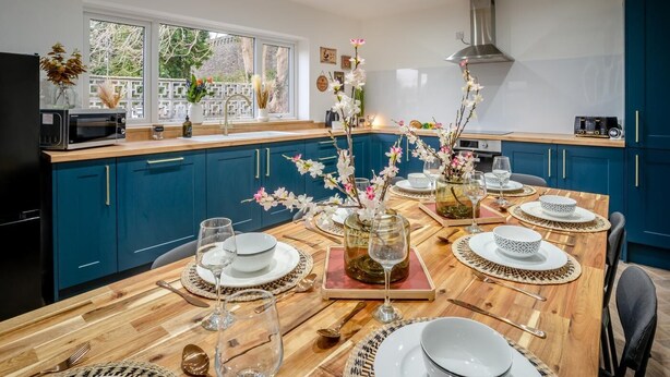 A modern kitchen with teal cabinets, a wooden dining table set for six, and a view of the outdoors through large windows. - 4 Bed in Brecon (oc-h30823) (Brecon)