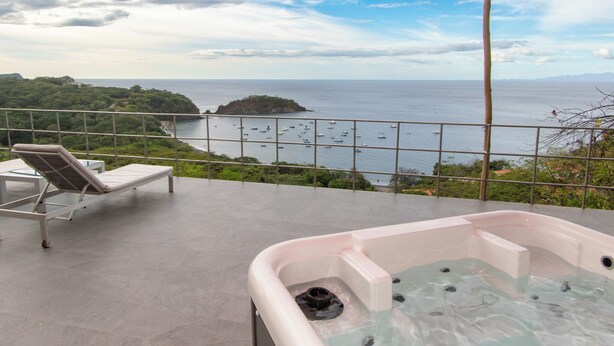 Balcón con jacuzzi y sillones de descanso con vistas a una masa de agua donde se ven barcos y una isla en la distancia.