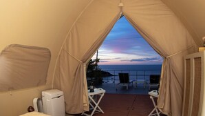 Habitación con carpa, una cama, una mesita y una silla. Se ve el océano y un atardecer.