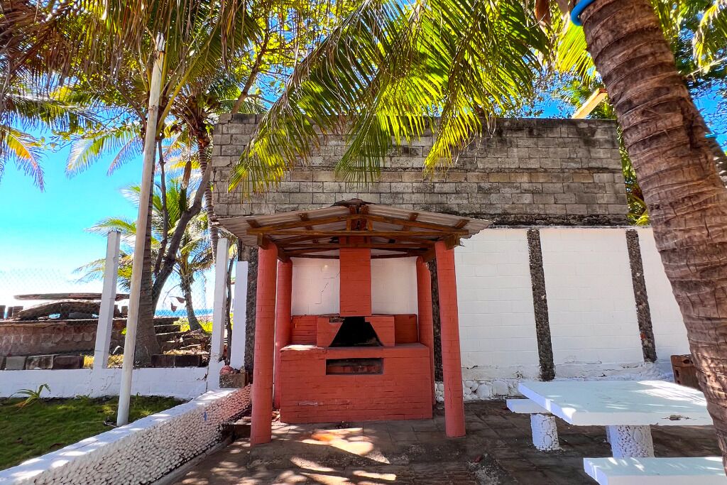 Um gazebo com base de tijolos e teto de madeira, rodeado por palmeiras e uma parede branca.