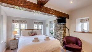 Un dormitorio con una cama grande, una pared de piedra, un televisor montado, una ventana con cortinas y una mesita de noche con lámpara.