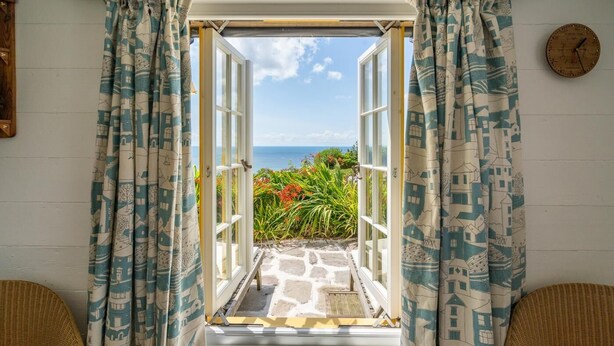 An open doorway leading to a balcony with a view of the sea, patterned curtains, and a clock on the wall. - 1 Bed in Millbrook (oc-b30672) (Tregonhawke)