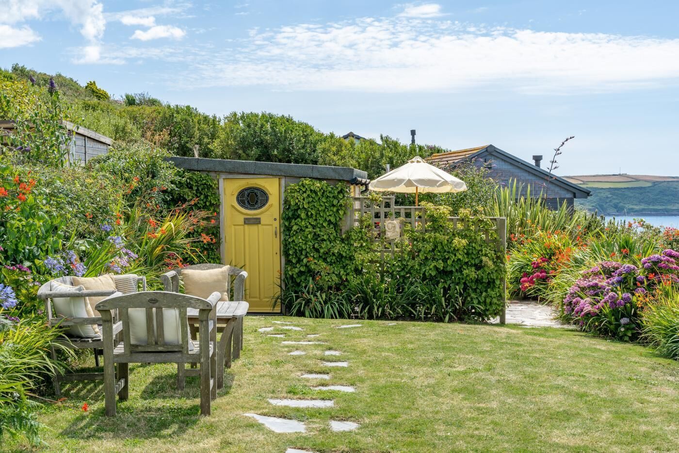 A garden with a yellow door, a wooden bench, and a stone path leading to a house with a view of the sea.