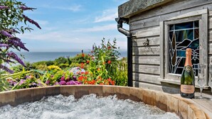 A wooden cabin with a hot tub filled with water, surrounded by colorful flowers and a view of the ocean. - 1 Bed in Millbrook (oc-b30672) (Tregonhawke)