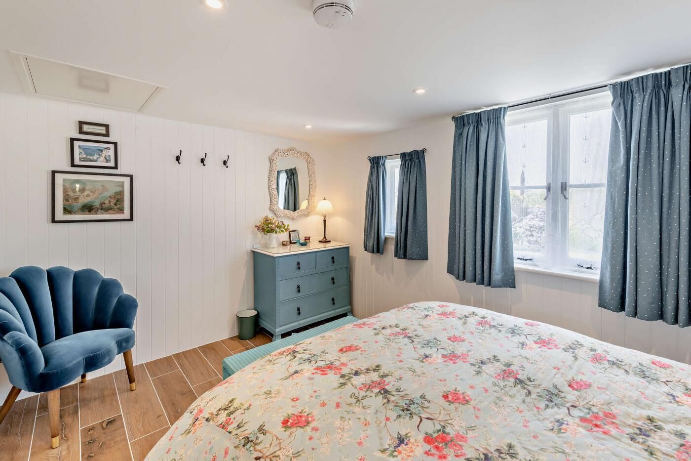 A bedroom with a floral bedspread, a blue armchair, a dresser with a mirror, and a window with curtains.