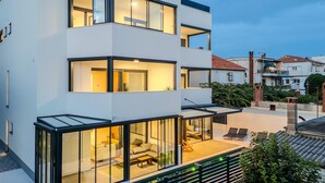 En moderne bygning med flere etager, store glasvinduer, en tagterrasse med udendørs siddepladser og et synligt poolområde.
