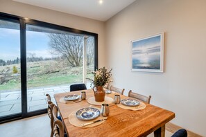 Een eetkamer met een groot raam, een houten tafel gedekt voor vier personen en een schilderij aan de muur.