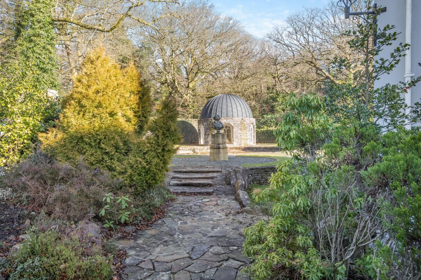 A stone pathway leading to a stone building with a dome roof, surrounded by dense foliage and trees.