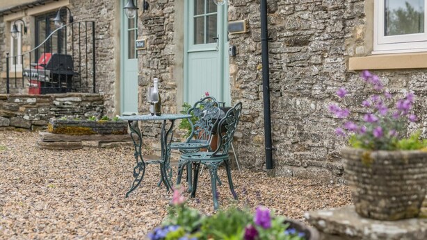 Un bâtiment en pierre avec une cour recouverte de gravier, une table et une chaise en métal, et un pot de fleurs contenant une plante aux fleurs violettes.