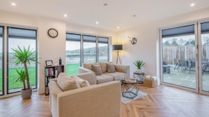 Ein großzügiges Wohnzimmer mit einer großen Couch, einem Couchtisch und einem Bücherregal. Große Fenster bieten einen Ausblick ins Freie.