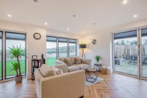 A spacious living room with a large sofa, a coffee table, and a bookshelf. There are large windows and a view of the outdoors. - 4 Bed in Pleasley Vale (oc-c30857) (Mansfield Woodhouse)