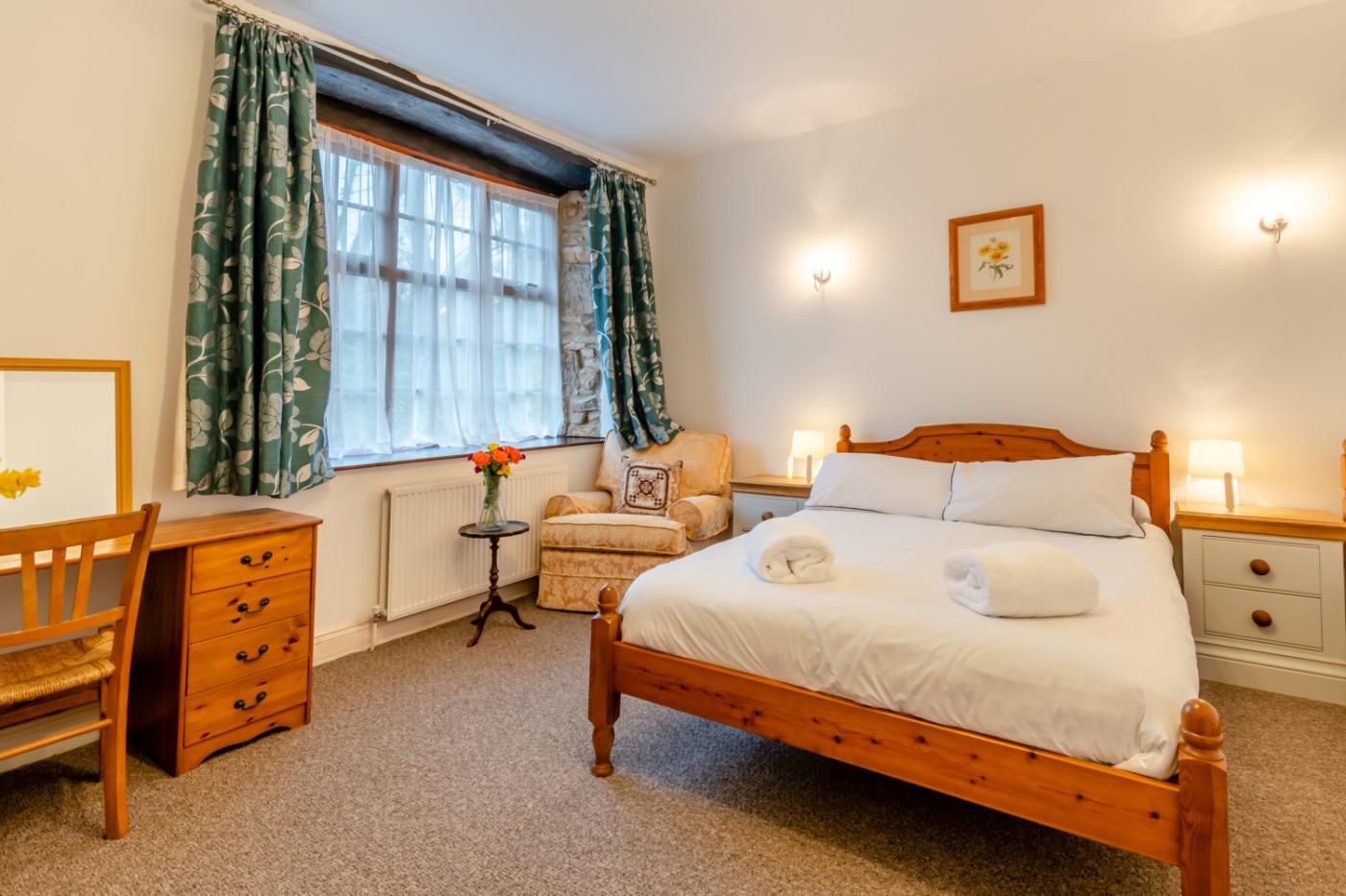 A bedroom with a wooden bed, a desk, a chair, a small table with flowers, and a radiator.