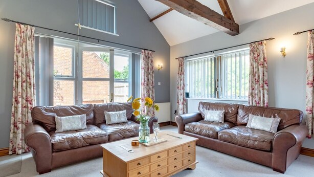 A spacious living room with two leather sofas, a wooden coffee table, and large windows with floral curtains. - 2 Bed in Horsham St. Faith (oc-b30838) (Horsham Saint Faith)