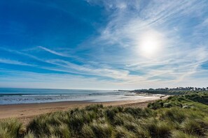 A coastal landscape with a beach, grassy dunes, and a distant shoreline with buildings. - 4 Bed in Seahouses (oc-s28973) (Seahouses)