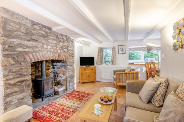 A cozy living room with a stone fireplace, a wooden sofa, a coffee table, and a view of the outdoors through a window. - 2 Bed in Kirkcudbright (oc-a30803) (Kirkcudbright)