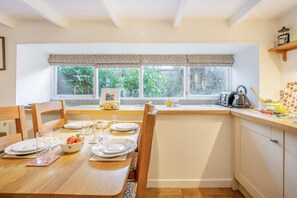 A dining area with a table set for two, a window with a view of greenery, and a kitchen counter with a kettle and a kettle pot. - 2 Bed in Kirkcudbright (oc-a30803) (Kirkcudbright)