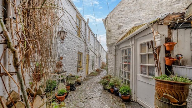 Une ruelle étroite pavée de pierres, bordée de bâtiments blancs, agrémentée de plantes en pot et ornée d’une statue.