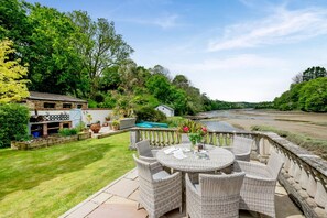 A patio with wicker furniture and a table, surrounded by a lush garden and a river view. - 2 Bed in Little Petherick (oc-o30788) (Little Petherick)
