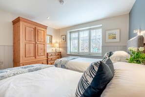 A bedroom with two beds, a wooden wardrobe, a window with blinds, and a wall-mounted lamp. - 2 Bed in Little Petherick (oc-o30788) (Little Petherick)
