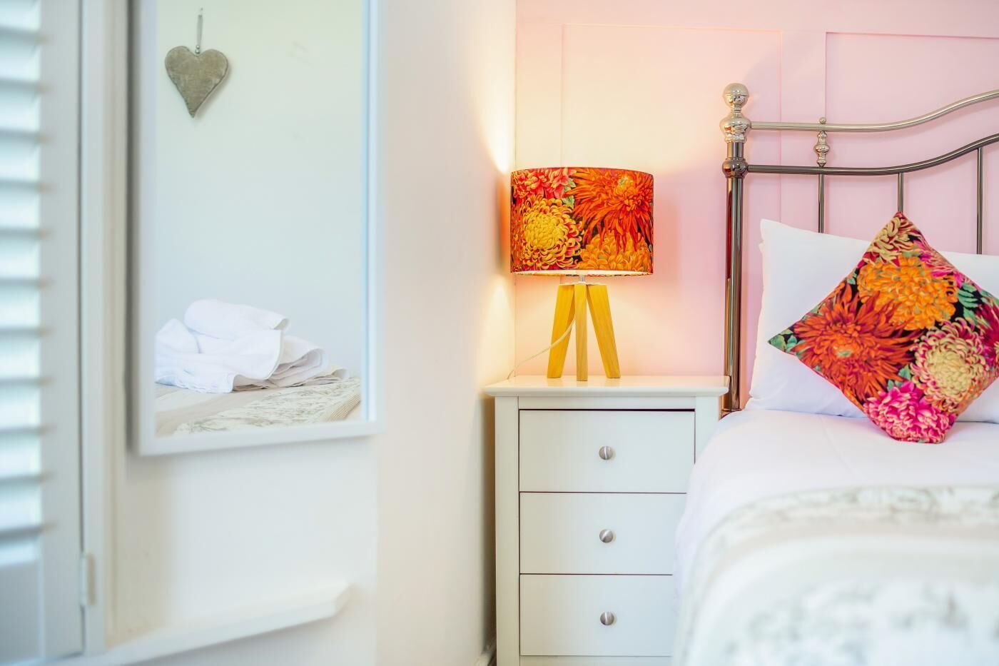 A bedroom with a bed, a bedside table with a lamp, a drawer, a framed picture on the wall, and a heart-shaped decoration.