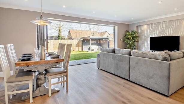 Een moderne woonkamer met een grijze bank, een eettafel met stoelen en een aan de muur bevestigde televisie.