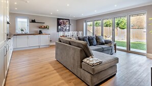 Een ruime woonkamer met een grijze bank, een keuken met witte kastjes en een grote schuifpui naar de achtertuin.