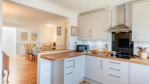 A modern kitchen with white cabinets, a wooden countertop, and a black stove. There is a dining area with wooden chairs and a table in the background. - 4 Bed in Ringstead (oc-k30839) (Ringstead)
