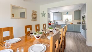 A dining area with a wooden table set for four, a kitchen in the background, and a star decoration on the wall. - 5 Bed in Walberswick (oc-m30723) (Walberswick)