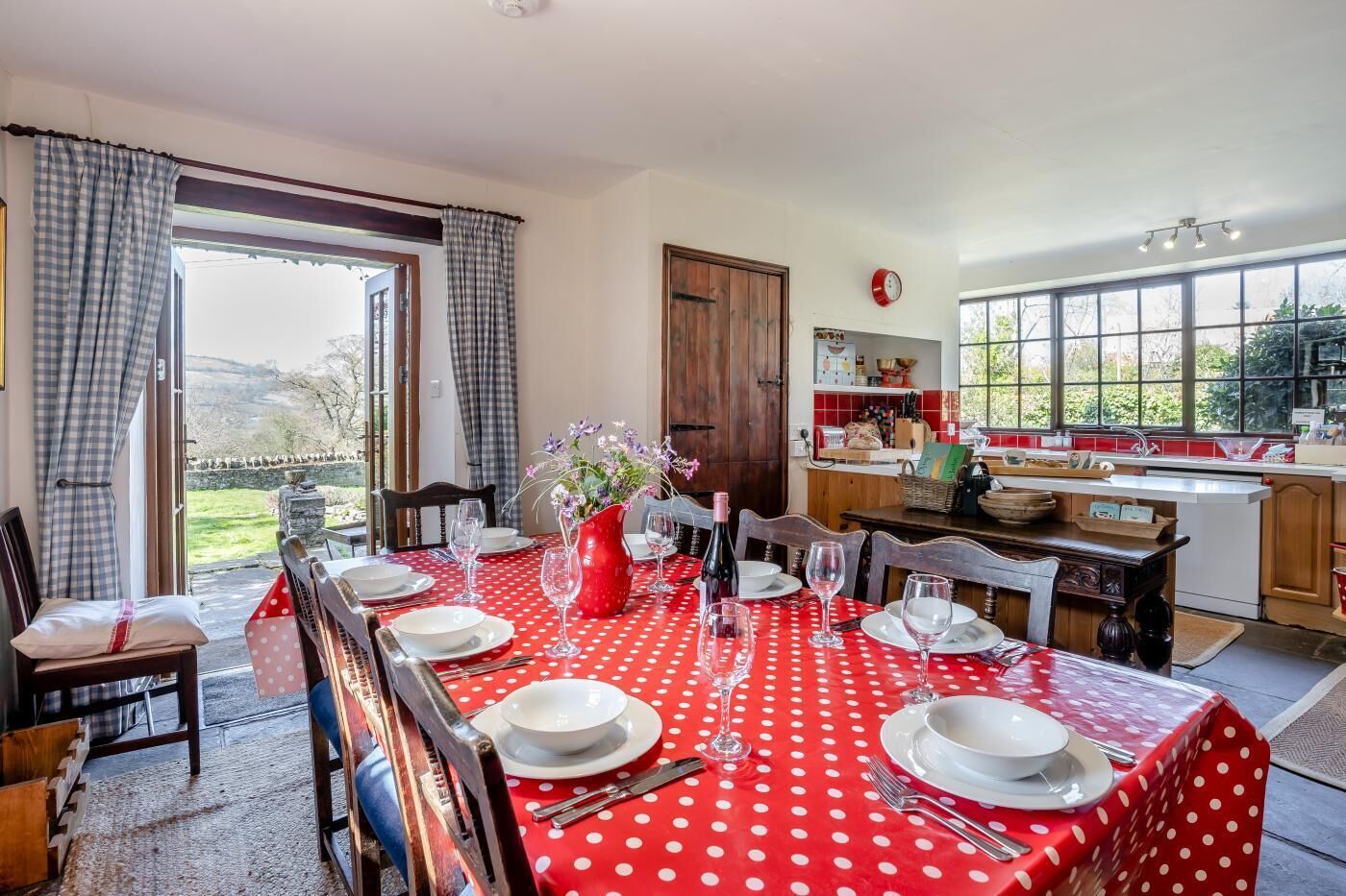 A dining area with a red polka dot tablecloth, wooden chairs, and a view of the garden through large windows.