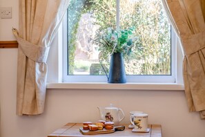 A window with curtains, a vase with greenery, a teapot, a mug, and pastries on a wooden table.