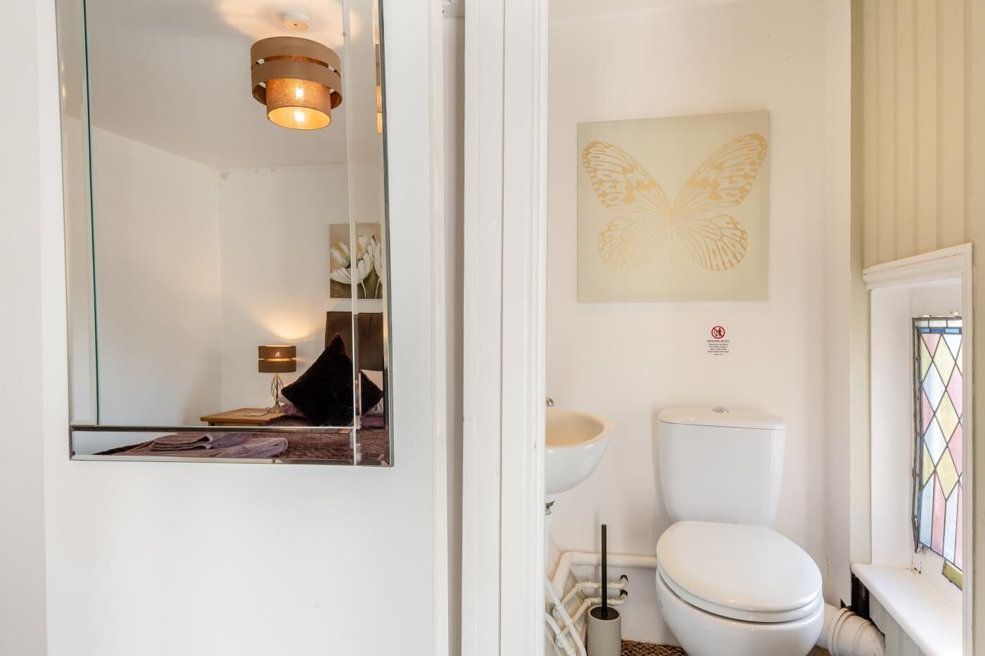 A modern bathroom with a toilet, sink, and a mirror reflecting a cozy room with a lamp and a chair.