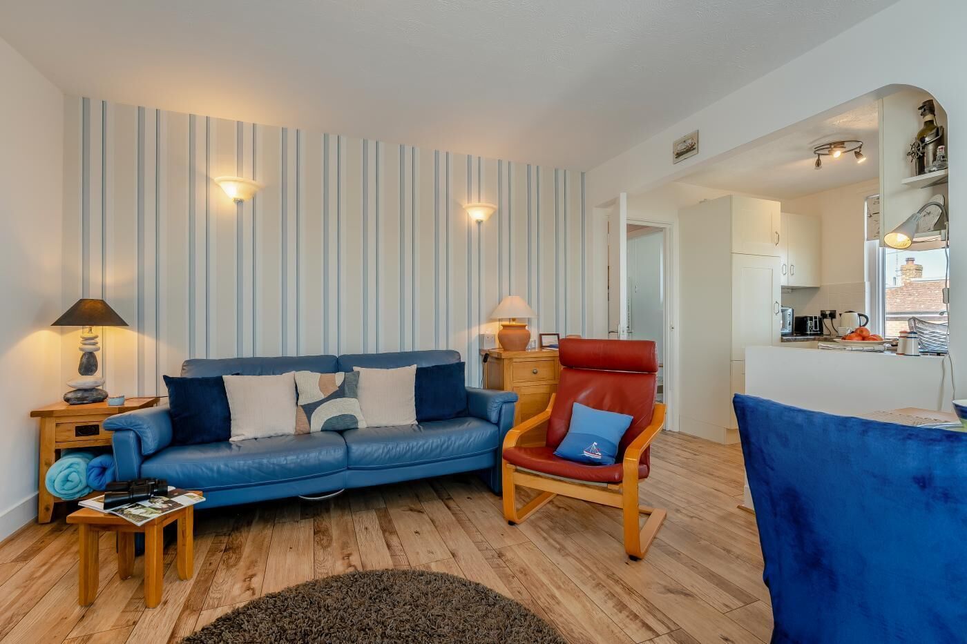 Un salón con un sofá azul, un sillón rojo y una mesa de centro de madera. Tiene papel tapiz a rayas y una alfombra pequeña en el suelo.