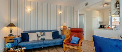 Un salón con un sofá azul, un sillón rojo y una mesa de centro de madera. Tiene papel tapiz a rayas y una alfombra pequeña en el suelo.
