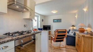 Une cuisine moderne équipée d’une cuisinière à gaz, d’un four et de meubles blancs. On y trouve une table à manger en bois avec des chaises, un canapé bleu et un téléviseur dans l’espace salon.