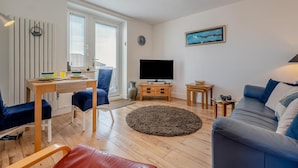 Un salon moderne comprenant un canapé, un téléviseur, un coin repas avec une table et des chaises, ainsi qu’une grande fenêtre équipée de stores.