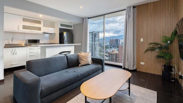 A modern living room with a grey sofa, a wooden coffee table, and a city view through large windows. - |EP| Nice Apt w/View, Balcony & Gym in El Poblado (Medellín)