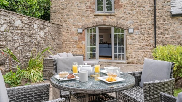 Edificio de piedra con zona de comedor que incluye mobiliario de mimbre y una mesa dispuesta con alimentos para el desayuno.