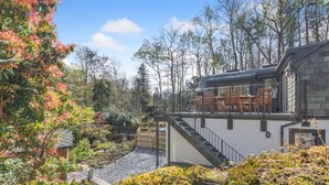 Une maison moderne dotée d’un balcon, de murs en pierre et d’un jardin où poussent différentes plantes et arbres.