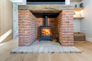 A wood-burning stove in a brick fireplace with a wooden beam above, a wicker basket, and a shelf with books. - 2 Bed in Moulton St. Mary (oc-c30849) (Moulton Saint Mary)