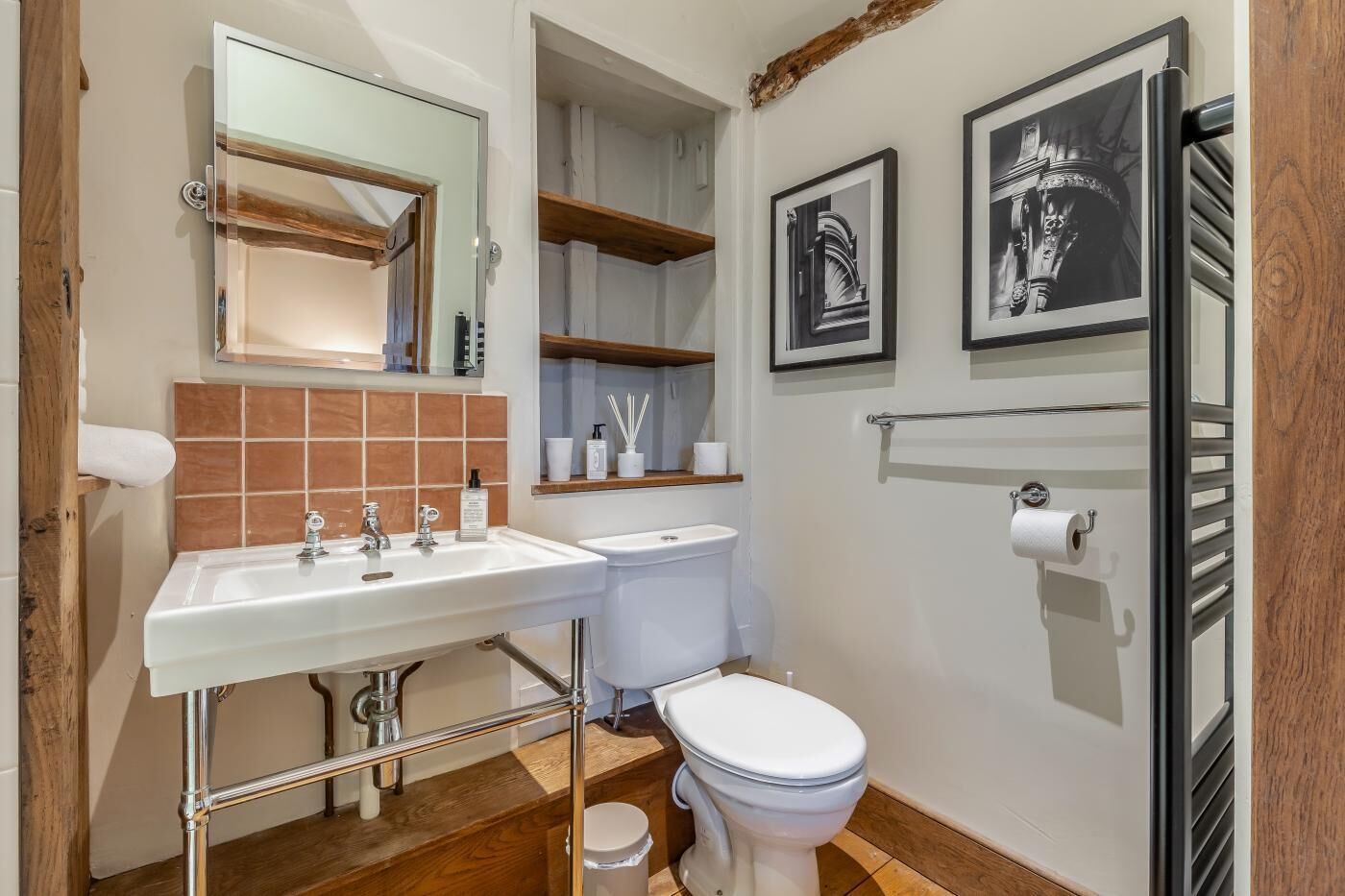 Une salle de bain avec un lavabo blanc, deux toilettes et un miroir. Il y a des étagères au-dessus du lavabo et un porte-rouleau de papier toilette fixé au mur.