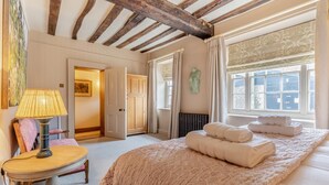 Quarto com cama grande, mesa de cabeceira com abajur, cadeira e vista para o exterior pela janela.