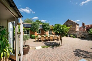A courtyard with a brick patio, a wooden table with chairs, and a stone building in the background. - 6 Bed in Walsingham (oc-r30797) (Little Walsingham)